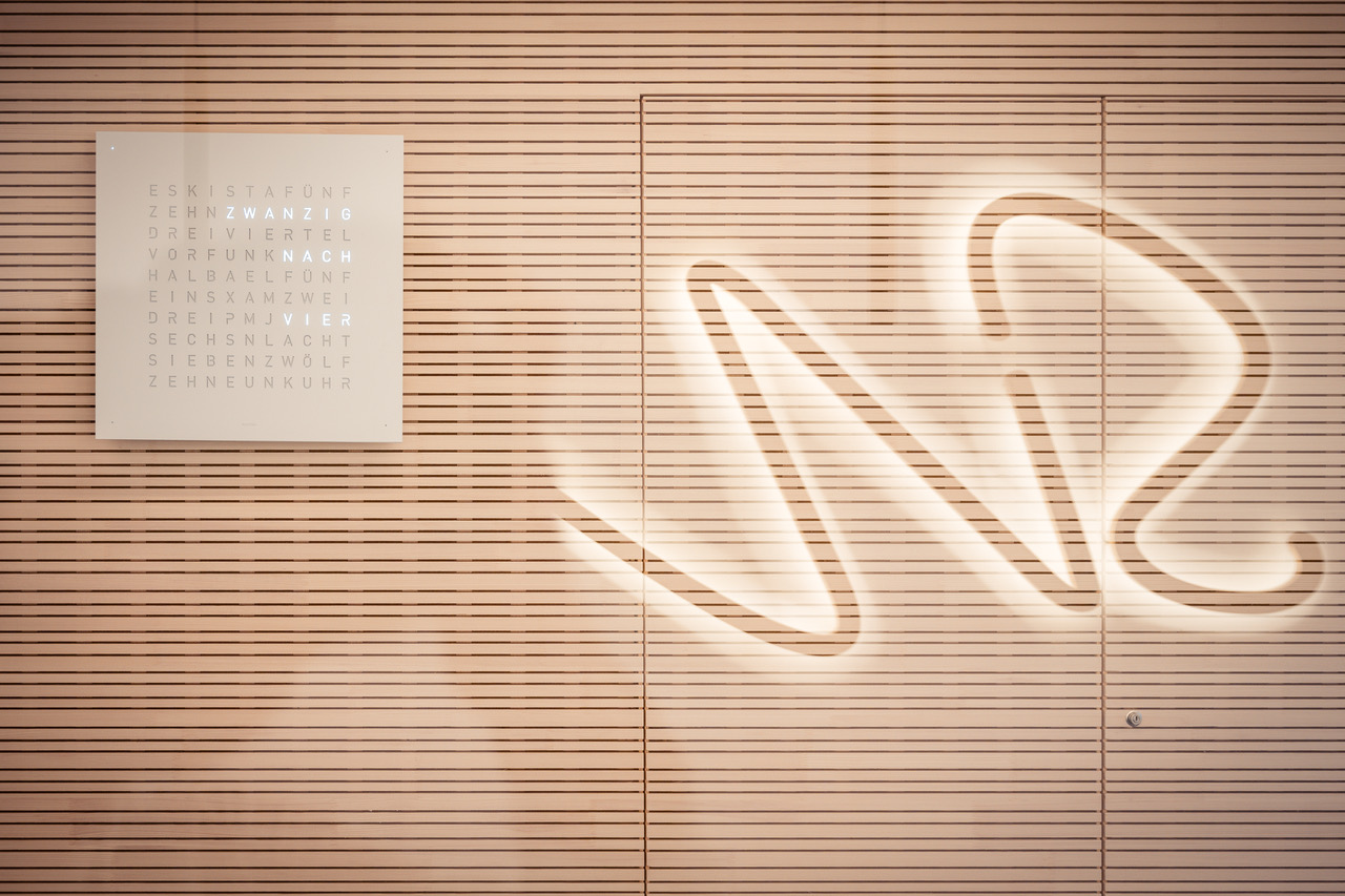 Zahnmedizin an der Lokhalle – zahnärztliche Kompetenz mit Herz 6 Holzlamellenwand mit Wortuhr und dem Schatten des beleuchteten Praxislogos.